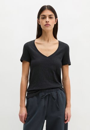 Woman standing against plain background wearing black V-neck short-sleeve shirt and dark drawstring pants, looking straight ahead.