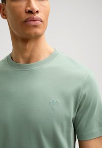 Kurzärmliges T-Shirt mit Rundhalsausschnitt aus leichtem grünen Baumwollstoff mit weicher Textur, das ein kleines gesticktes Logo auf der Brust hat.