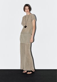 Modella in un outfit a strisce beige si trova su un pavimento nero, indossando una lunga gonna e una maglietta a maniche corte, abbinata a una collana con ciondolo nero.