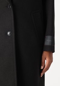 Svart ullblandingsfrakk med dobbelspent design, store knapper og et merke på armen med røde sømdetaljer.