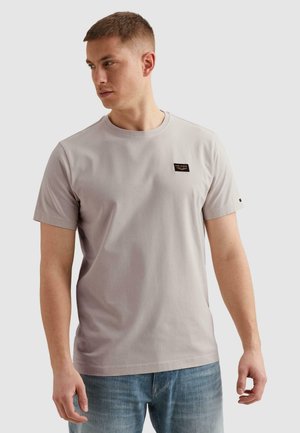 Młody mężczyzna ubrany w jasnoszary T-shirt z krótkim rękawem z małą czarną naszywką z logo na piersi, patrzący w lewo na jednolitym tle.