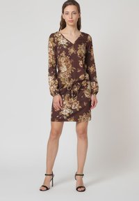 Vestido floral marrón con mangas largas, escote en V y cintura con cordón. Presenta un patrón botánico en tonos crema y verde. Sandalias de tacón tipo cuerda.