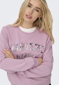 Lichtroze sweatshirt met de tekst "HARVEST MARKET" in bloemenpatronen. Ontworpen met een ronde hals en geribbelde mouwen, zachte textuur.
