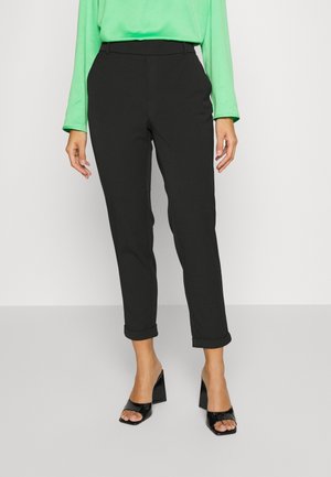 Vrouw draagt zwarte slim-fit cropped broek met omgeslagen pijpen, een felgroene lange mouw top en zwarte open teen blokhakken, staand op een grijze vloer.