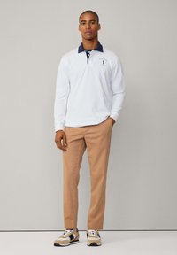 Valkoinen pitkähihainen polo paita, jossa on tummansininen kaulus ja logo, yhdistettynä vaaleanruskeisiin housuihin ja beigeihin lenkkikengiin, joissa on mustia yksityiskohtia.