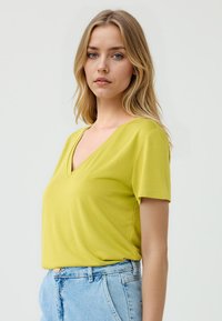 Donna con capelli biondi mossi indossa una maglietta a maniche corte color verde lime e pantaloni shorts in denim a vita alta, in piedi contro uno sfondo neutro.