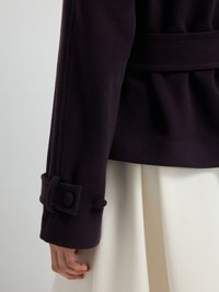 Il cappotto in misto lana viola presenta una silhouette strutturata, dettagli a fiocco ai polsini e una vita con cintura. Una gonna bianca spunta al di sotto.