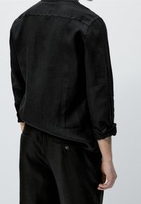 Man in een zwart shirt met lange mouwen en opgerolde mouwen, en zwarte broek, gezien van achteren tegen een effen achtergrond.