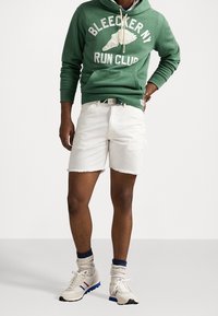 Grön hoodie med trycket "BLEECKER NY RUN CLUB", vita denimshorts med fransar, vita sneakers med blå och röda accenter, och ribbade strumpor.