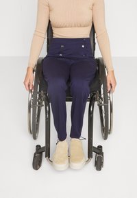 Navy trousers with faux buttons, beige ribbed top, and light tan shoes. Subject seated in a wheelchair with metal wheels and sleek design.