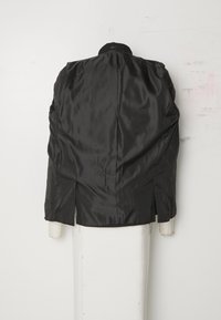 Giacca nera in satin con una texture liscia, caratterizzata da un colletto alto, tasche laterali e spacchi sul fondo per facilitare il movimento.