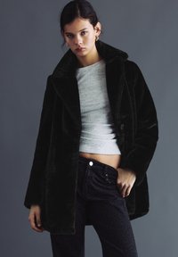 Tânără purtând o haină neagră din blană ecologică peste un top gri scurt și pantaloni închiși la culoare cu model, stând pe un fundal gri simplu.