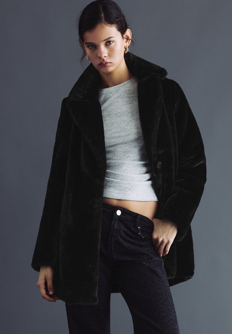 Ung kvinde iført en sort faux pelsjakke over en grå croppet top og mørke mønstrede bukser, stående mod en ensfarvet grå baggrund.