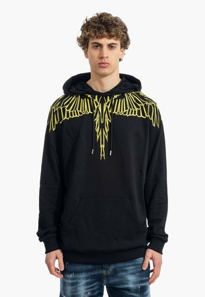 Jeune homme aux cheveux bouclés portant un sweat à capuche noir avec des motifs jaunes en forme d'ailes sur les épaules et le haut de la poitrine.