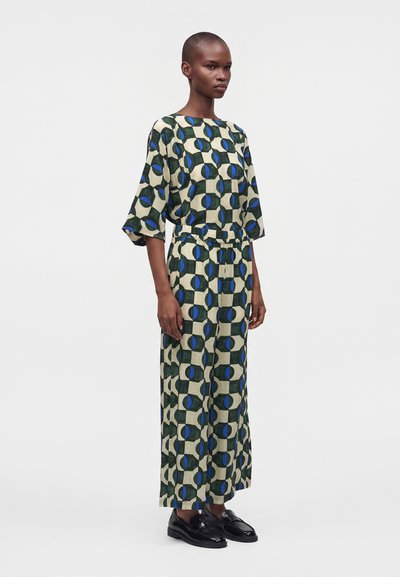Modèle portant un haut ample assorti et un pantalon large avec un motif géométrique bleu et vert, debout sur un fond uni.