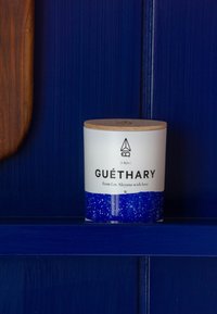 Vit cylindrisk ljushållare med en blå glittrig botten och lock av trä. Har texten "GUÉTHARY" på en mörkblå bakgrund.