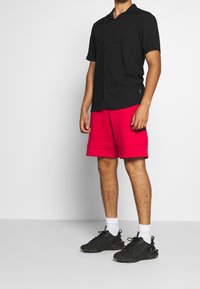 Schwarzes Kurzarm-Hemd mit Knöpfen und einer Brusttasche, kombiniert mit roten Shorts und schwarzen Sneakern. Weiße Knöchellocken sind sichtbar.