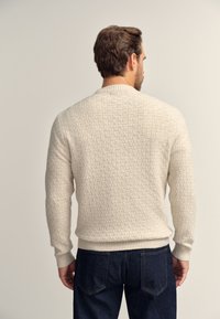 Ljusbeige texturerad tröja med ribbad krage och muddar, med ett rutmönster. Bärs med mörkblå jeans.