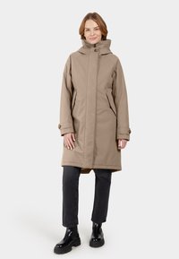 Beige lange jas met een hoge kraag, knoopsluiting en zijzakken, gecombineerd met zwarte jeans en stevige zwarte laarzen. Gladde stoftextuur.