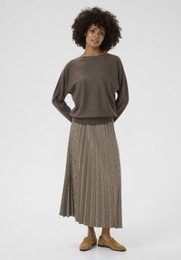 Brūns, garām piedurknēm, pārāk liels džemperis, savienots ar plisētu, līdz ceļiem, rūtainu svārku. Kājās gaišas krāsas loferi, neitrāls fons.