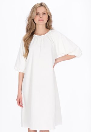 Blondýna stojící s rukou na boku, oblečená v volných bílých šatech s balónovými třičtvrtečními rukávy a nařaseným výstřihem.