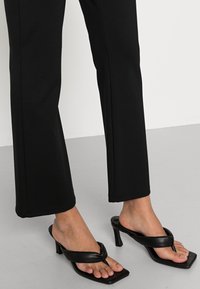 Czarne sandały na obcasie z designem palca, z gładką skórzaną fakturą i kwadratowym noskiem. Spodnie są czarne o prostym kroju.