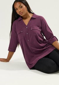 Camicia a maniche lunghe di colore viola scuro con due tasche sul petto, dettagli in metallo argentato e polsini arrotolati, abbinata a pantaloni neri.