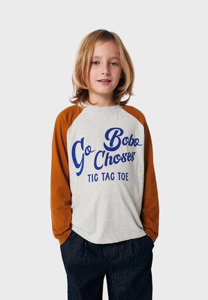 Graues Langarmshirt mit braunen Raglanärmeln, versehen mit blauer Schrift, die "Go Bobo Choses Tic Tac Toe" lautet. Gestreifte dunkle Hose.