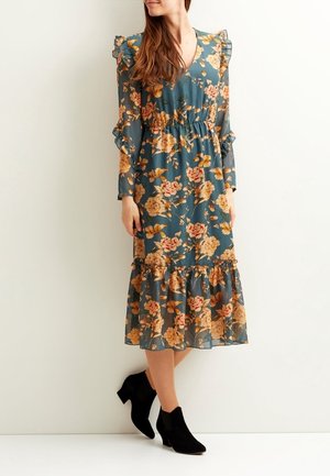 Teal värinen kukkamekko, jossa on röyhelöiset hihat, kireä vyötärö ja kerroksellinen helma. Koristeena on suuria lämpimiä kukkia. Mustat nilkkasaappaat.