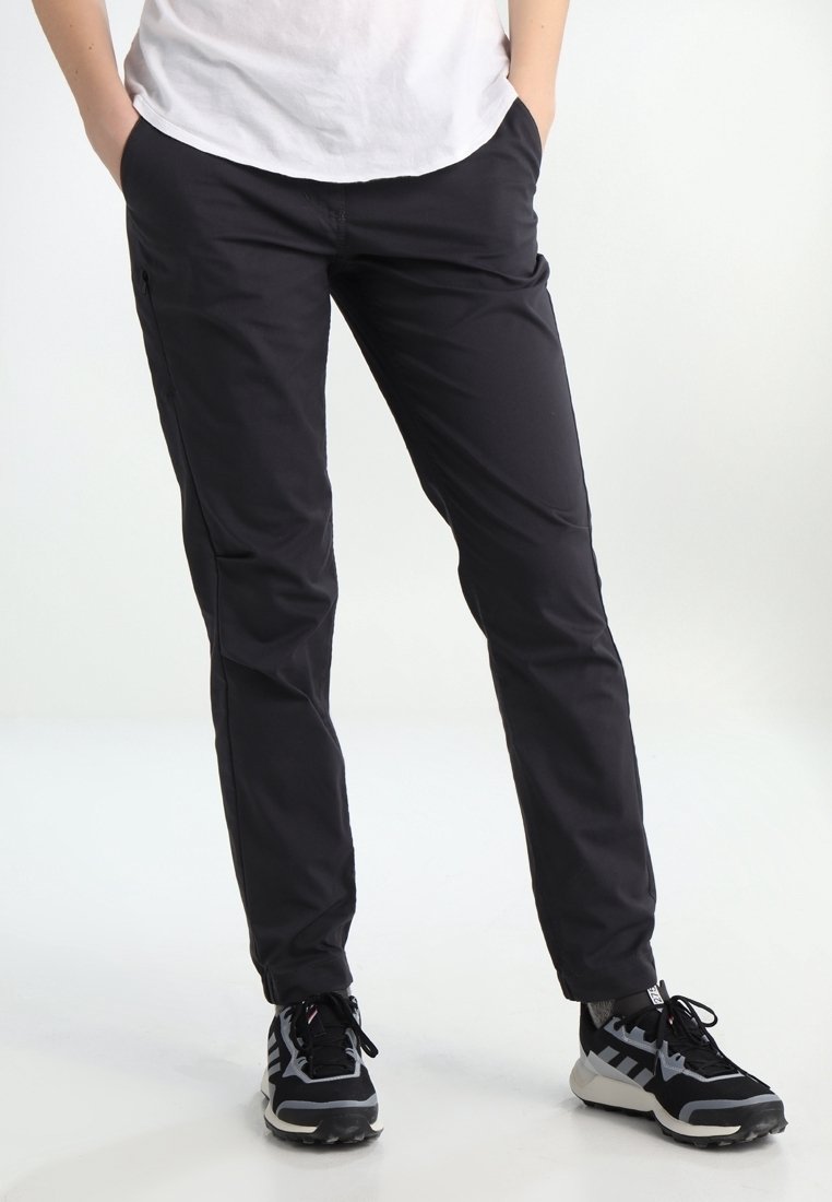 Personne portant un pantalon noir et des baskets noires et grises, les mains dans les poches, debout sur un fond blanc.