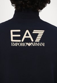 Tumesinise kõrge kaelusega dressipluus. Seljal valge trükitud logo "EA7 EMPORIO ARMANI". Pehme tekstuur, klassikaline disain ja vabaaja lõige.