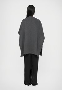 Sivi vuneni pončo prekriva figuru, s otvorenim izrezom i opuštenim rubom. Crne široke hlače dovršavaju odjevnu kombinaciju.