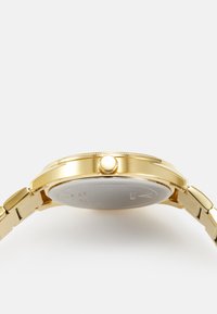 Reloj de tono dorado con caja redonda, bisel texturizado y pulsera lisa. Presenta una corona lateral y una parte posterior de caja de color plateado.
