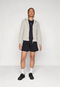 Világosszürke cipzáras dzseki tengerészkék póló felett, fekete rövidnadrággal és fekete tornacipővel párosítva. A fehér zoknik a cipők felett láthatók.