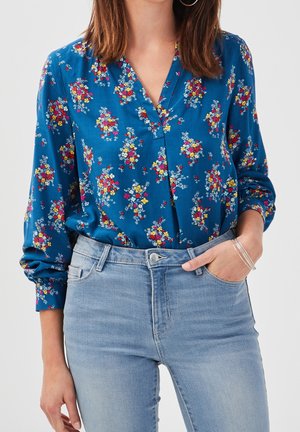 Femme portant un chemisier bleu à fleurs à manches longues, rentré dans un jean taille haute bleu clair, la main dans la poche et des bracelets argentés au poignet.