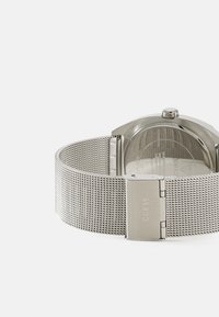 Reloj de acero inoxidable con correa de malla, con una caja redonda, corona y un cierre con "GUESS" grabado. La parte trasera es transparente.