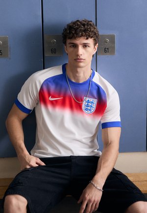La maglia Nike presenta un tessuto bianco con una sfumatura di rosso e blu, colletto e maniche blu, e un emblema dell'Inghilterra. Indossata con pantaloni corti, posizionata in uno spogliatoio.