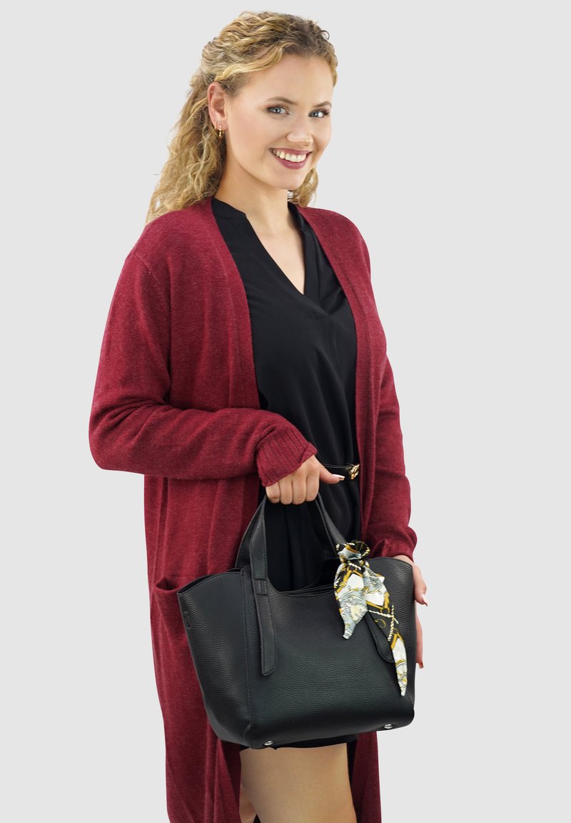 Zwarte leren handtas met een textuur, voorzien van twee handvatten en een decoratieve zijden sjaal die aan een handvat is geknoopt. Model draagt een bordeauxrode cardigan.