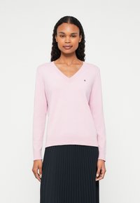 Maglione rosa chiaro a scollo a V realizzato in tessuto morbido, dotato di maniche lunghe e un piccolo logo del marchio sul lato sinistro del petto. Indossato con una gonna nera plissettata.