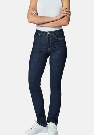 Person som har på seg mørkeblå slim-fit jeans, hvit ermeløs topp og hvite joggesko, armer krysset over brystet mot ensfarget bakgrunn.