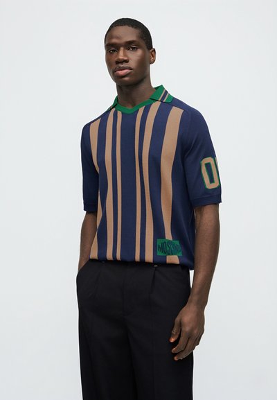 Άνδρας που φοράει μπλε σκούρο και μπεζ ριγέ πόλο με πράσινο γιακά και μαύρο παντελόνι, στέκεται μπροστά σε απλό φωτεινό φόντο.