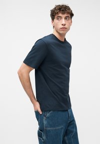 Νέος άνδρας με σγουρά μαλλιά, που φορά μπλε σκούρο t-shirt και φαρδιά τζιν παντελόνια cargo, στέκεται με ένα χέρι στην τσέπη.