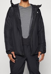 Giacca nera isolata con zip frontale e cappuccio regolabile, indossata sopra una camicia grigia. Presenta polsini in velcro e tasche laterali.
