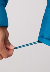 Blue insulated jacket with a textured fabric. Hand pulling a drawstring at the sleeve cuff, featuring a contrasting dark blue adjuster.