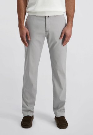 Man die lichtgrijze broek met rechte pijpen draagt, wit overhemd in de broek gestopt, en donkerbruine instapschoenen, staande tegen een effen achtergrond.