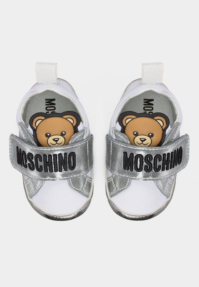 Λευκά και ασημένια παπουτσάκια μωρού με πρόσωπα αρκουδάκια στη γλώσσα και την επιγραφή "MOSCHINO" στις ασημένιες λουράκια που διασχίζουν το πάνω μέρος.