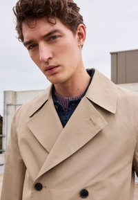 Trench coat beige de doble botonadura con solapas muesca, grandes botones negros y una textura suave. Lleva puesto un jersey de punto oscuro.