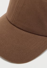 Καφέ καπέλο από καμβά με δομημένο γείσο, εμφανείς ραφές και υφή στην επιφάνεια. Η σχεδίαση έχει στερεωμένη κορυφή χωρίς ραφές και καθαρές ακμές.