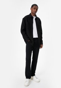 Camisa negra de botones con dos bolsillos en el pecho, combinada con pantalones negros y zapatillas blancas, con un corte relajado y una textura suave.