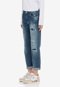 Mujer que lleva puestos unos jeans azules desgastados con puños, una blusa blanca de manga larga y zapatillas blancas con detalles en beige y texto negro en las suelas.